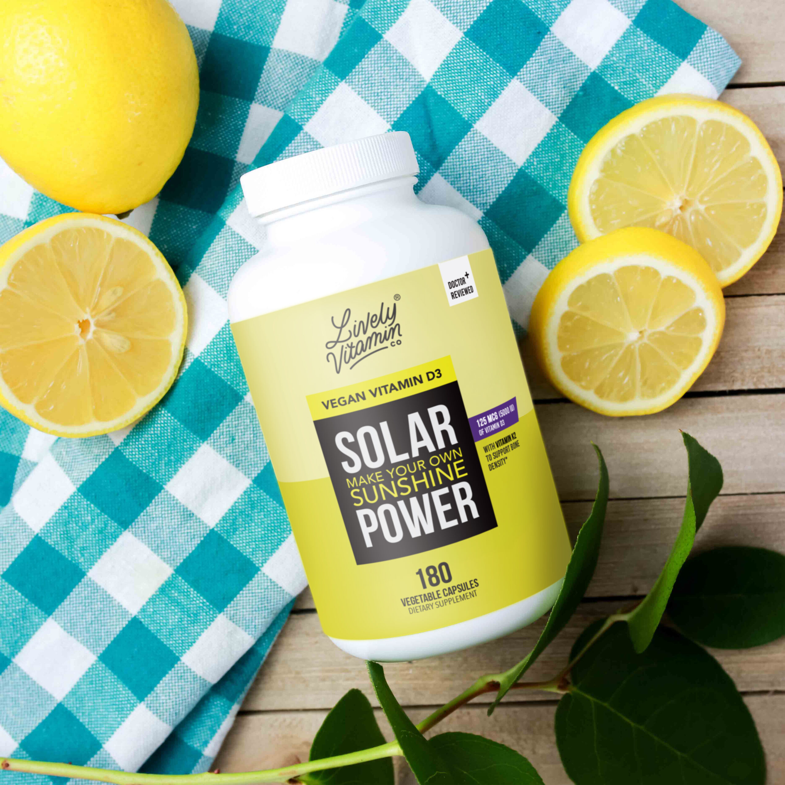 A white bottle of Lively Vitamin Co.'s Solar Vegan Vitamin D3 supplement rests on a turquoise checkered cloth, surrounded by fresh lemons and green leaves. The label highlights "Make Your Own Sunshine Power" in bold text, with the supplement containing 125 mcg (5000 IU) of vitamin D3 in 180 vegetable capsules.