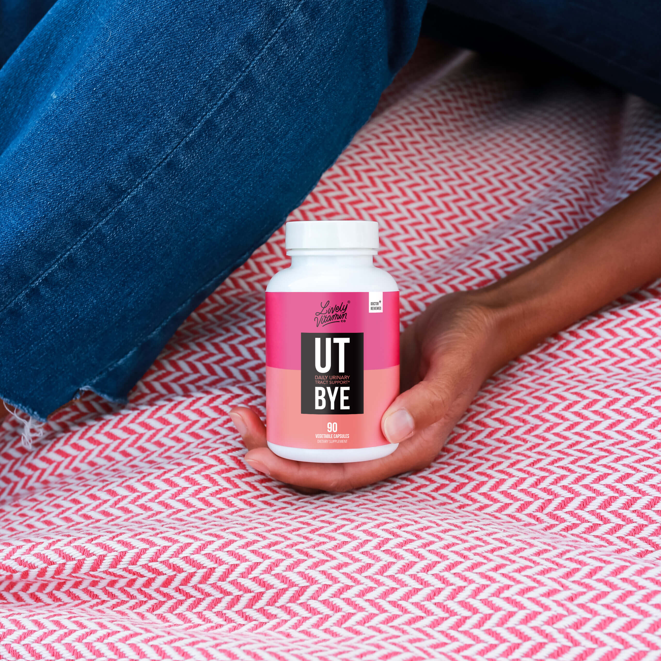 MockUpUT-Bye A person holding a white bottle of "UT Bye" dietary supplement by Lively Vitamin Co., designed for daily urinary tract support. The label features a bold pink and coral design, states “Doctor Reviewed,” and indicates the bottle contains 90 vegetable capsules. The person is seated on a red and white herringbone-patterned fabric with denim visible.